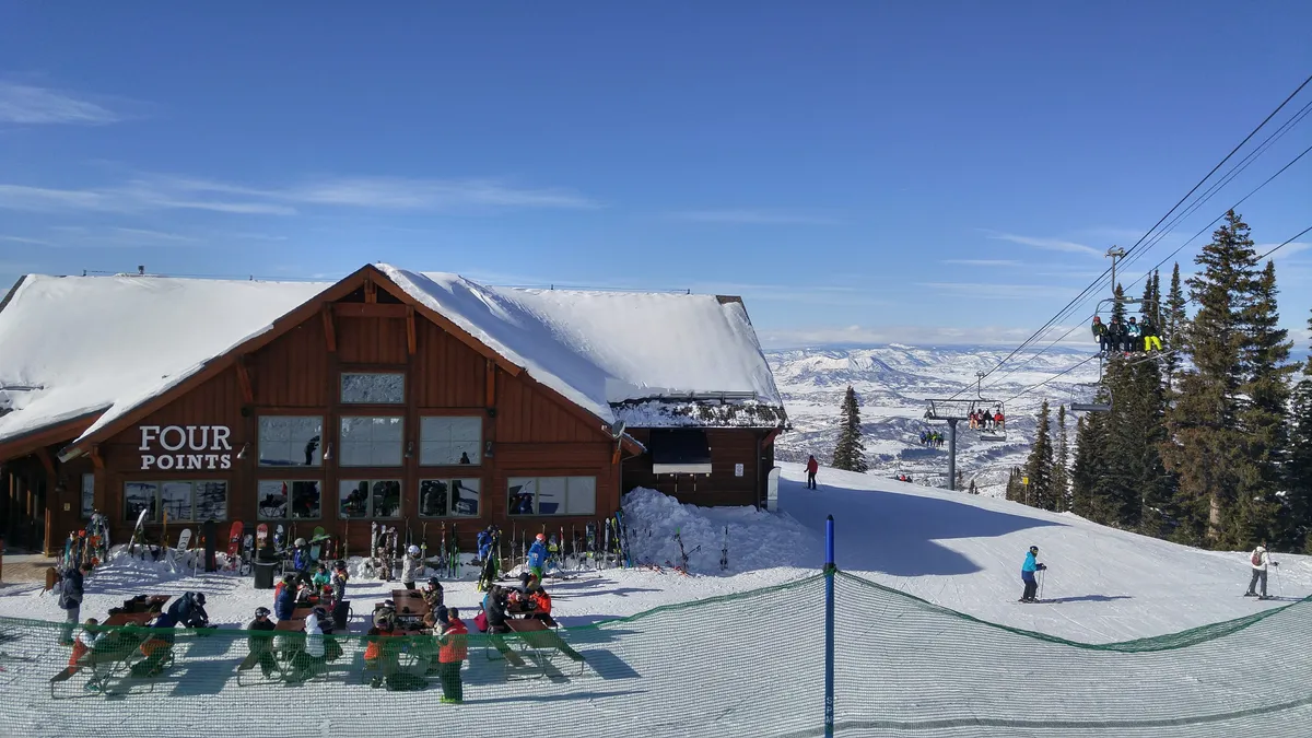 This lively exterior shot captures the main ski lodge building at Steamboat Ski Resort, with people skiing and snowboarding near the ski lift. The outdoor seating area is filled with guests enjoying food, drinks, and the vibrant mountain atmosphere. Snow-covered peaks and blue skies set the scene for an energetic winter adventure.