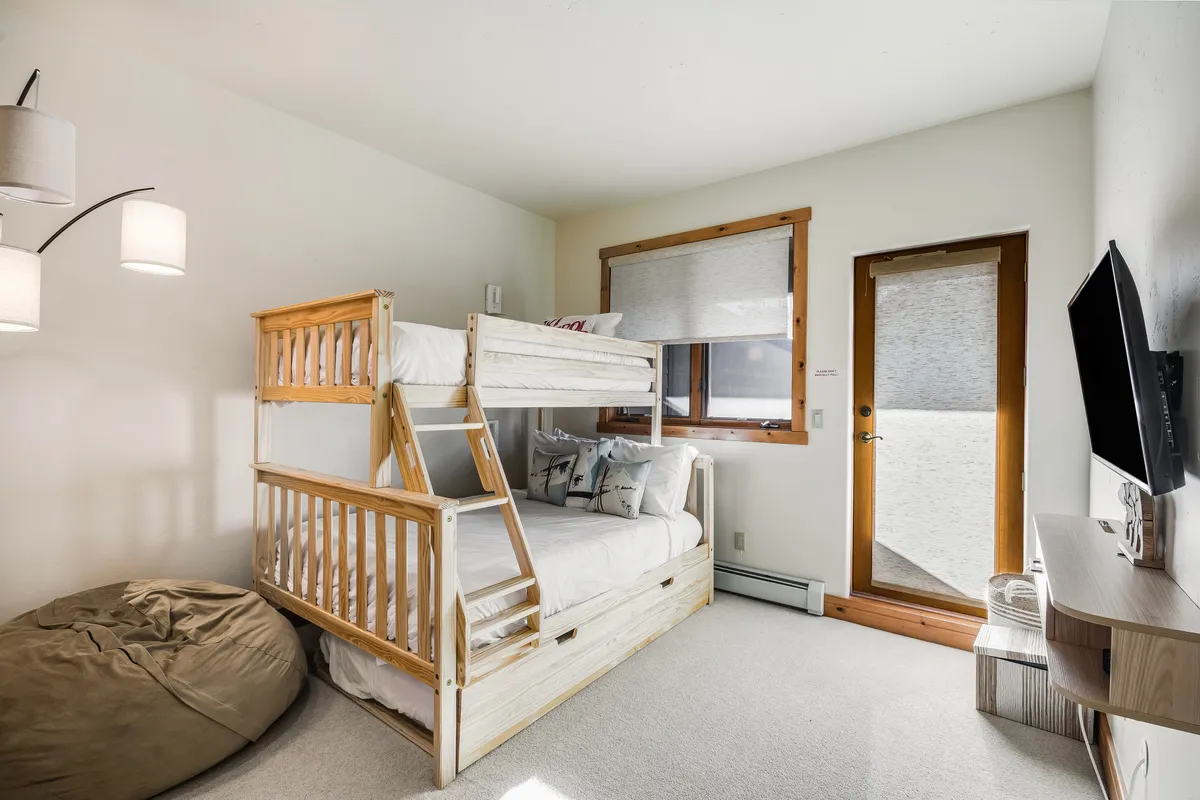 A playful and comfortable bunk room with a sturdy bunk bed, a cozy bean bag chair, a window filling the space with natural light, and a television mounted on the wall—designed with children or groups in mind.