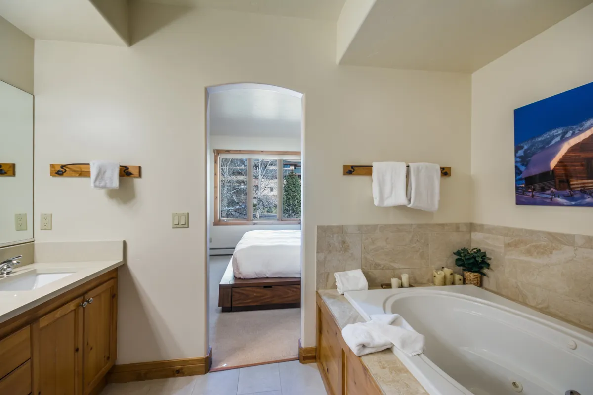 A tranquil bathroom scene with a soaking bathtub by a large window, complemented by a vanity mirror and natural light. The space is designed for relaxation and self-care, with views out to nature enhancing the calming atmosphere.