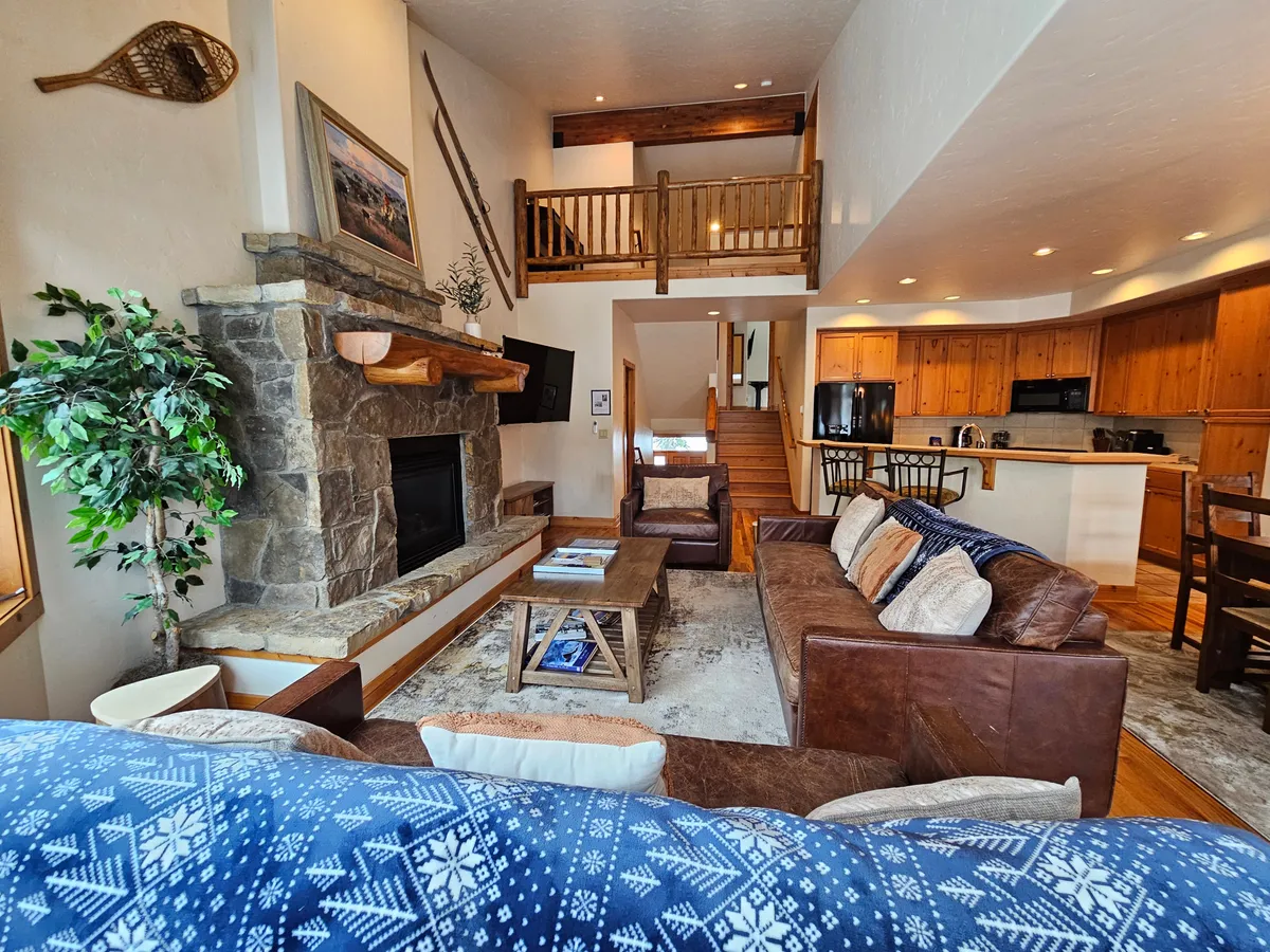 A warm, open-concept living area featuring a cozy fireplace, inviting sofa, visible wooden stairs, a kitchen area in the background, and decorative artifacts. The space is arranged for both relaxation and entertaining, suggesting a comfortable gathering spot after a day of mountain activities.