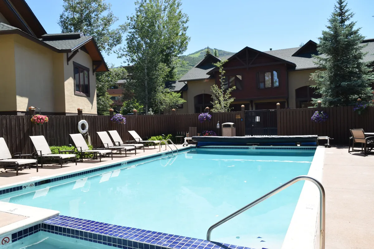 A tranquil outdoor pool area framed by leafy trees and comfortable lounge chairs, with buildings visible in the background. The space is designed for relaxation and community events, offering scenic sunset views.