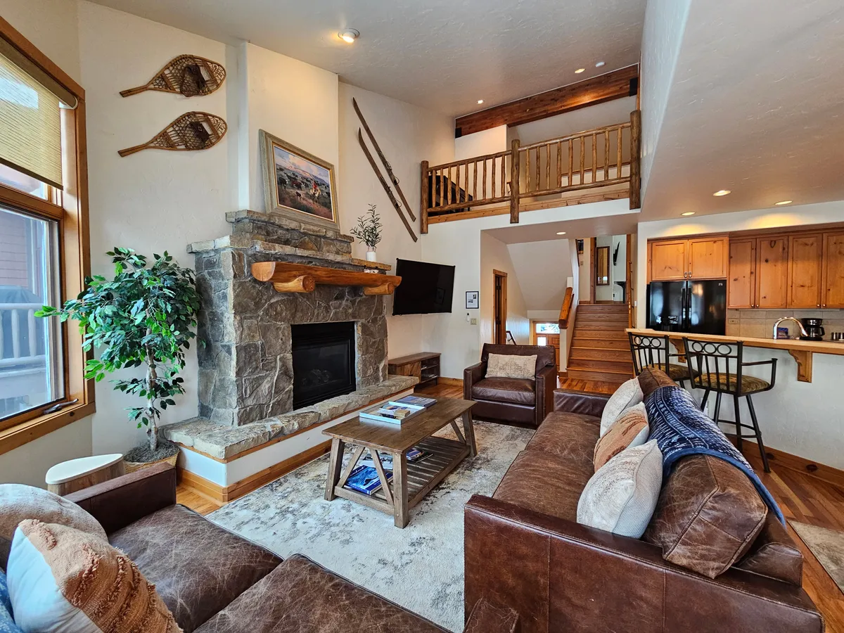 This image features a cozy living area centered around a beautiful stone fireplace, with a comfortable seating arrangement and a view of the wooden staircase. The room is softly lit and invites relaxation, evoking a sense of warmth and tranquility.