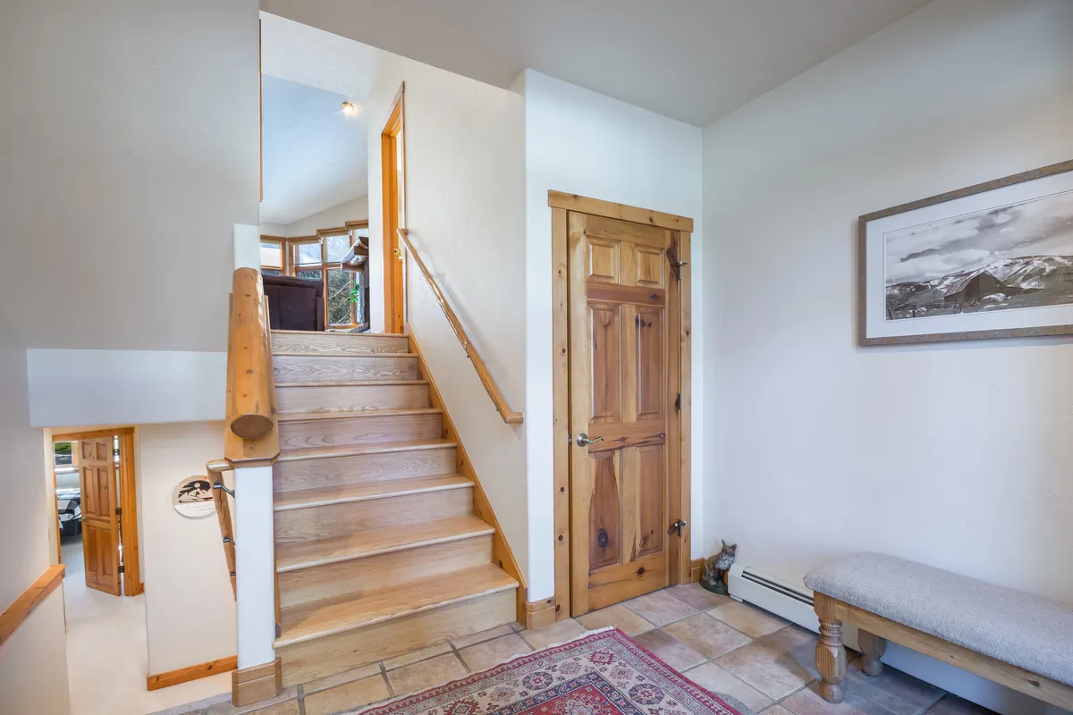This image captures an inviting entryway with a wooden staircase, a bench for sitting or removing boots, a wooden door hinting at direct access, and a framed picture on the wall. The space is practical and organized, evoking the atmosphere of a mudroom designed for easy transitions in and out of the home.