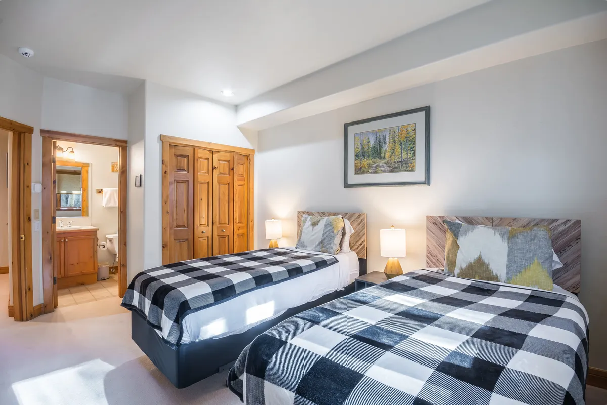 A bright twin bedroom with two plaid bedding twin beds, warm wooden closet doors, framed landscape art, and soft bedside lighting, all set in a comfortable, relaxed environment.