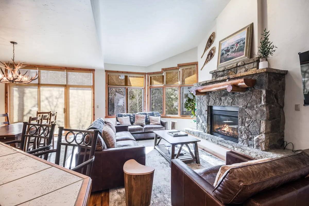 A cozy, sunlit interior highlighting a comfortable seating area by the fireplace, large windows that flood the space with light, and a stylish dining table set for communal meals. The setting exudes warmth and is ideal for both relaxing by the fire and enjoying group feasts.