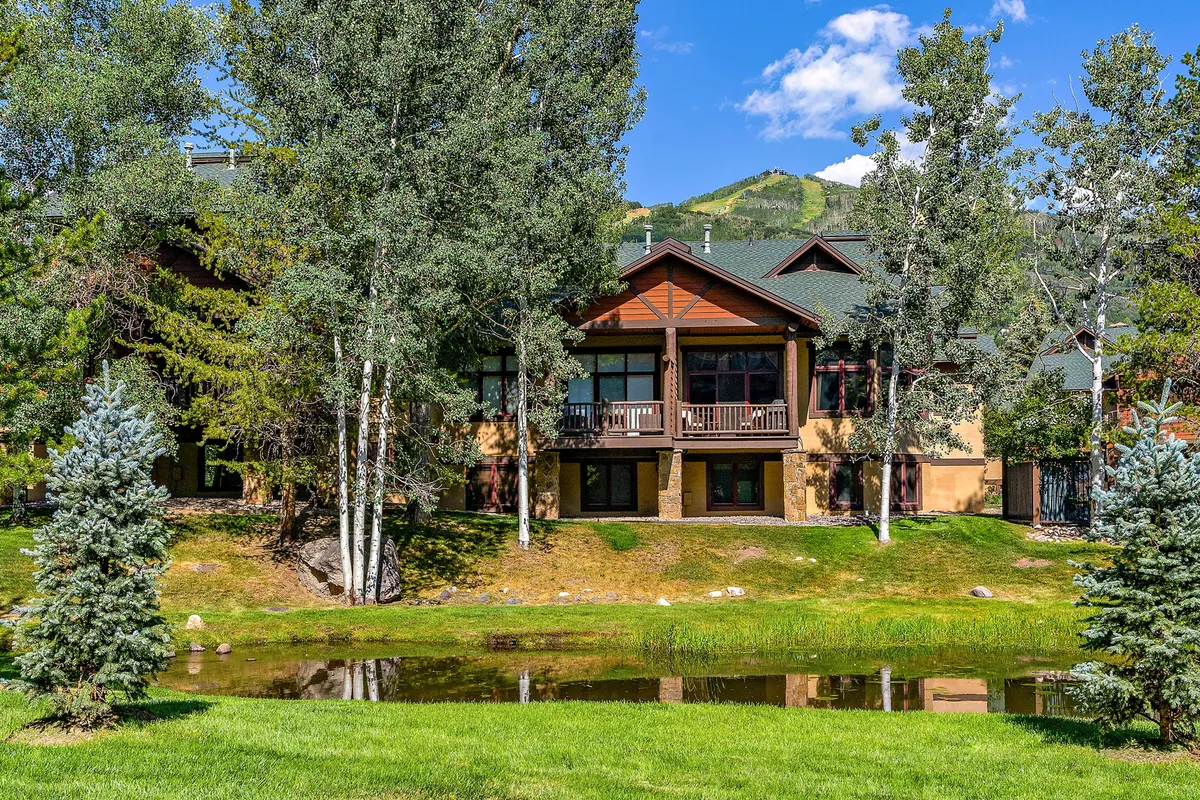 The scene features the exterior of the property, a tranquil pond, and surrounding trees. Reflections play across the water at sunset, and the natural landscaping hints at opportunities for wildlife viewing and serene moments by the water.