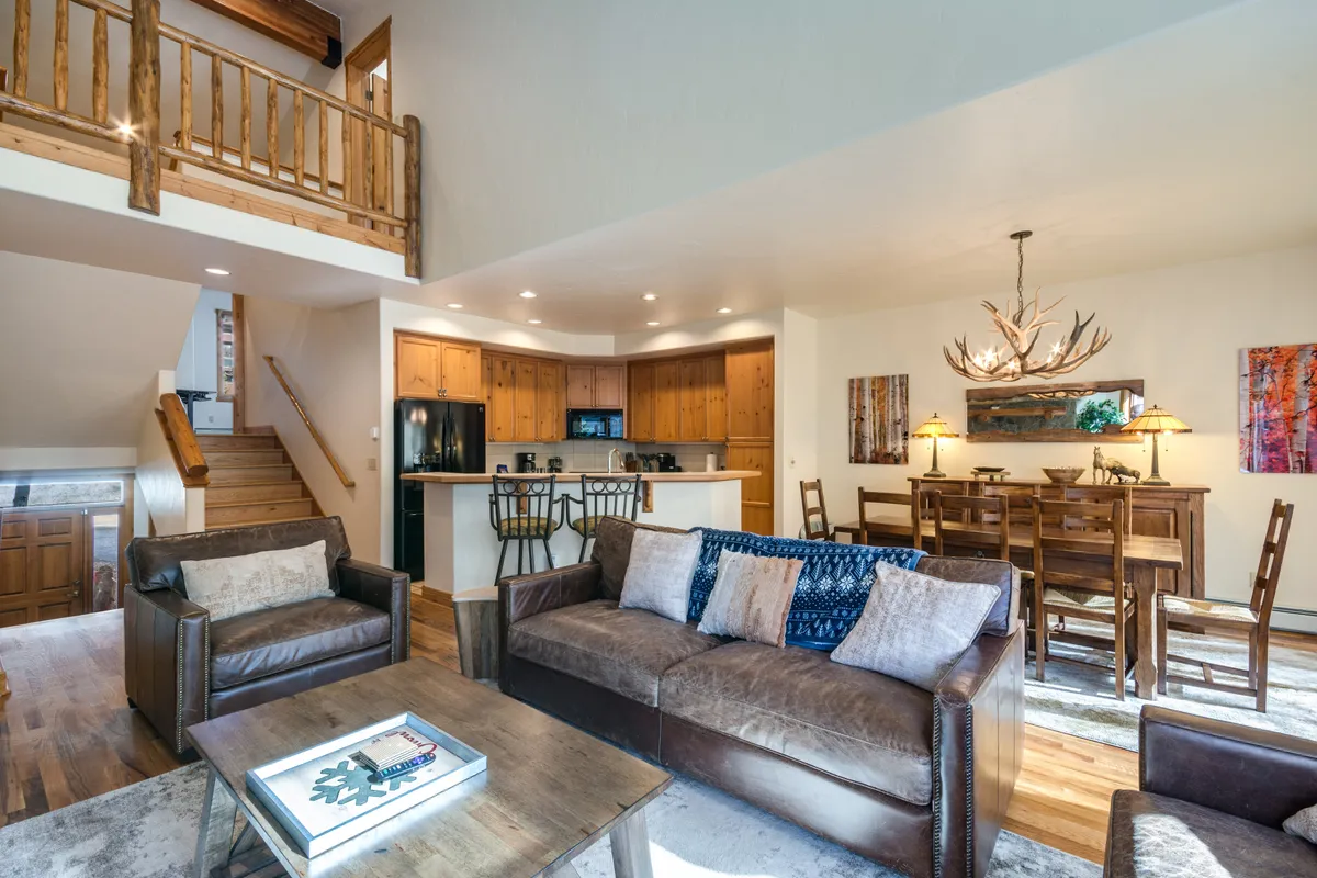 A welcoming view of the living and dining area, featuring a comfortable sofa with decorative pillows, a substantial dining table under an antler chandelier, and a glimpse of the kitchen’s rustic wooden cabinetry. The layout is inviting and designed for both entertaining and relaxation.