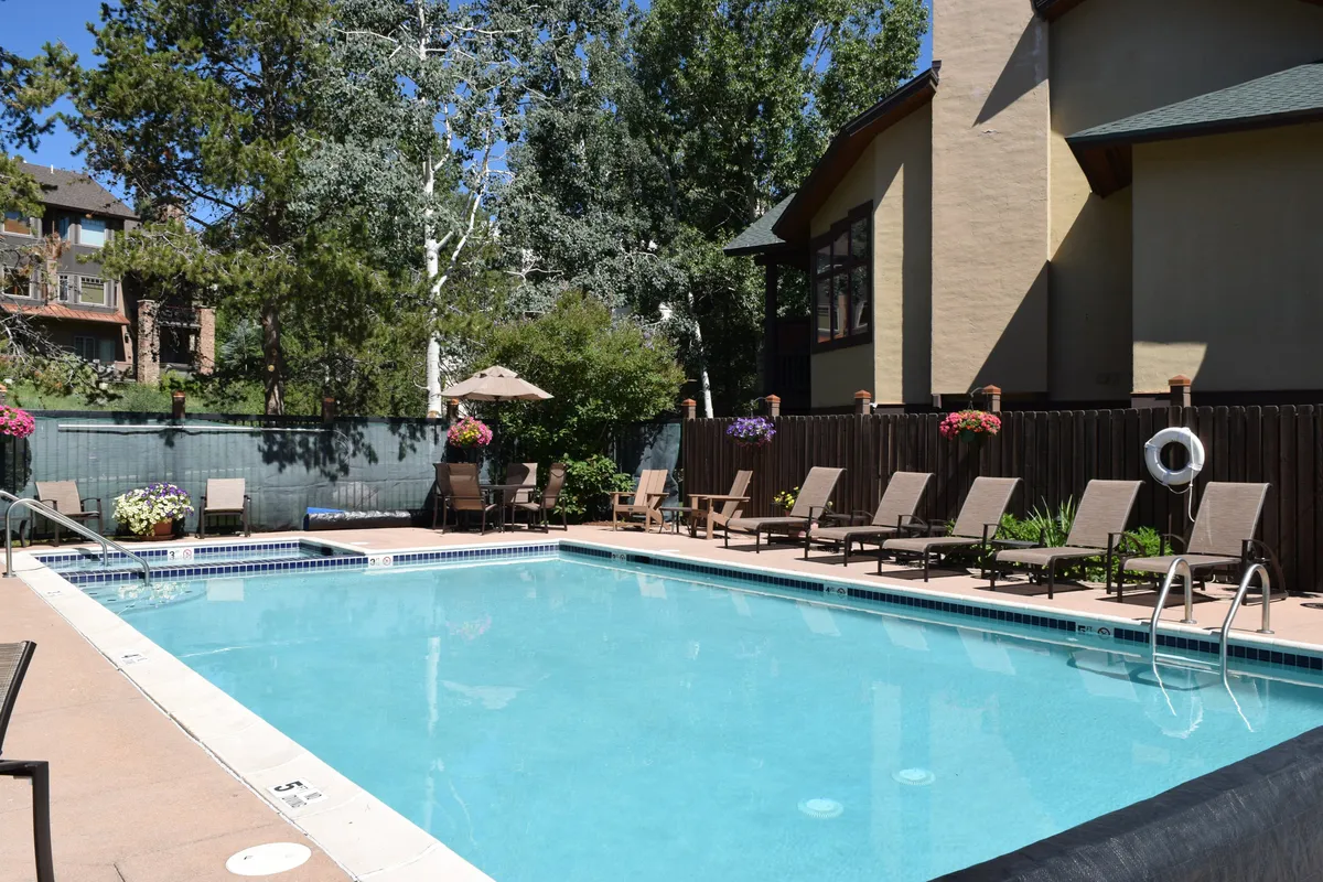 A sunlit swimming pool is surrounded by bright lounge chairs, flower pots, and umbrellas, creating a cheerful and welcoming space for both relaxation and social gatherings. The setting is outdoors, with room for multiple guests to enjoy the amenities together.