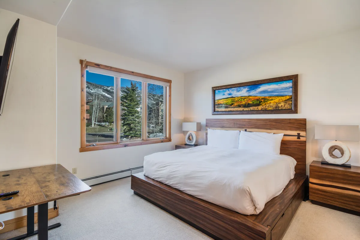 This image showcases a large bed with inviting bedding, a window providing natural light and a scenic view, artwork above the headboard, and wooden furniture, creating a restful and serene atmosphere ideal for a primary or king bedroom suite.