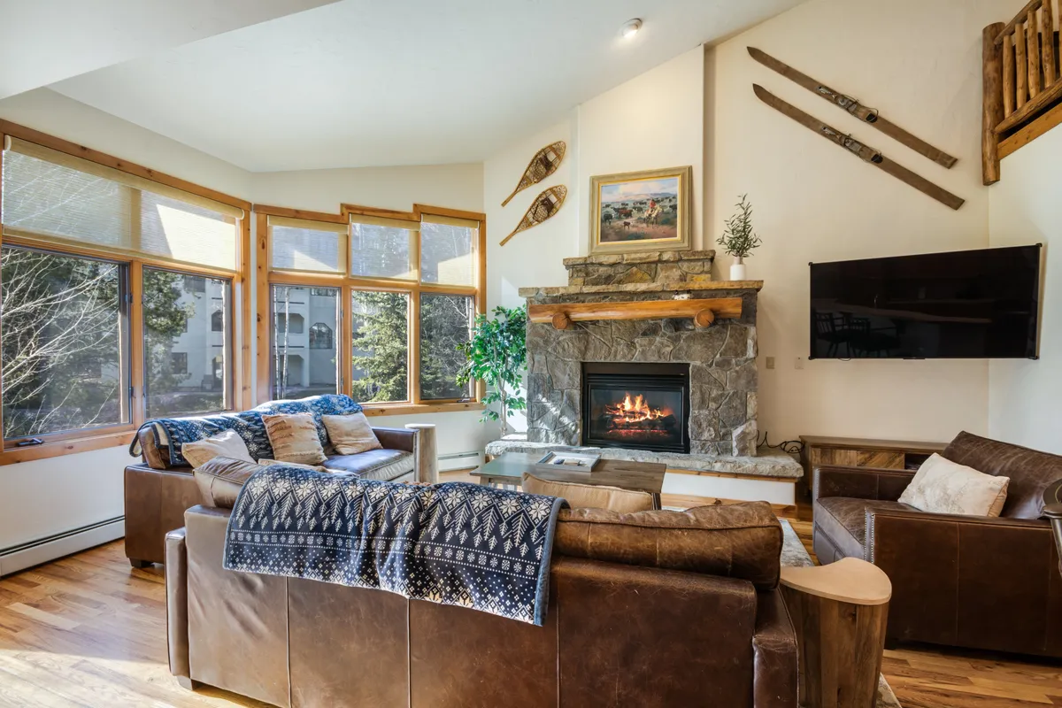 The image shows a spacious and inviting living room centered around a cozy gas fireplace. Large windows frame views of the snowy outdoors, while plush seating and decorative ski accents create a warm, mountain-inspired atmosphere. The room is perfect for relaxing after a day on the slopes, with the glow of the fire and soft lighting enhancing the welcoming mood.